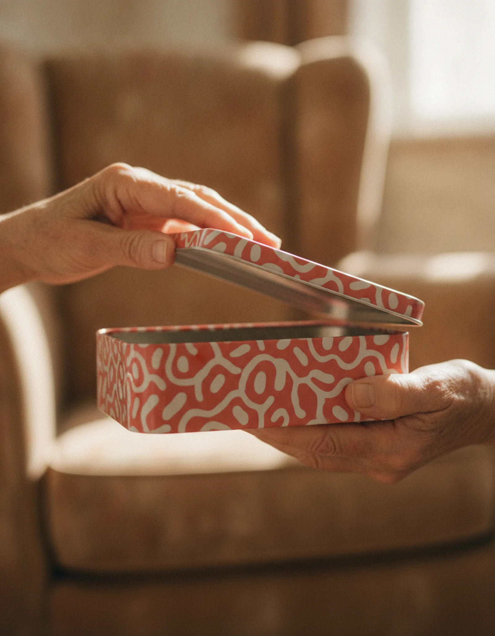 Older adult's hands opening a rectangular hinged-lid tin one-handed, thumb on the lip