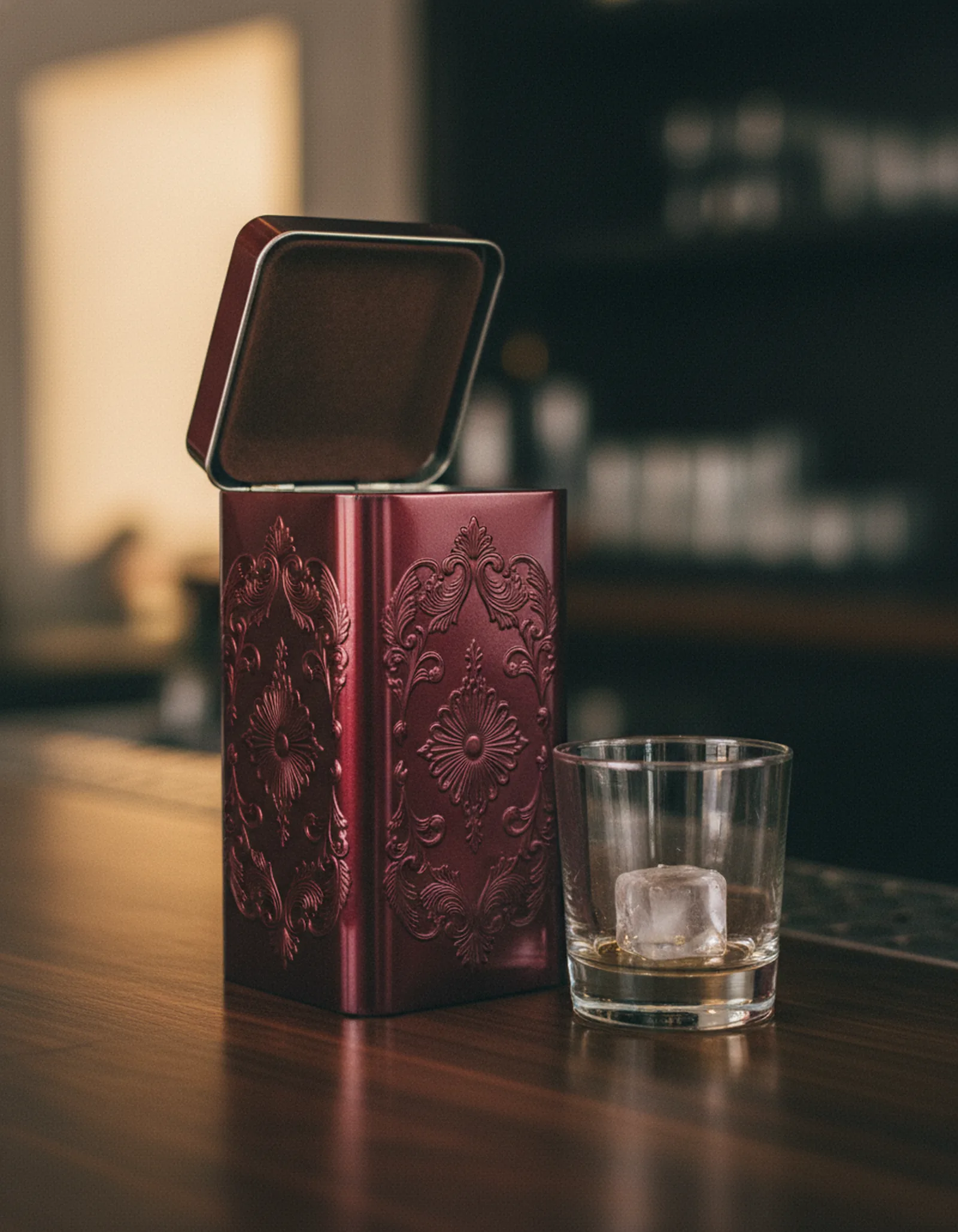Premium burgundy and gold spirits gift tin on a dark bar surface with a tumbler beside it